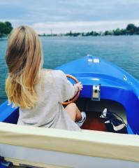 Woman with blonde hair steering a blue boat on a lake, wearing a light gray sweater, with a bag on the seat next to her, overcast day, calm water, scenic view, adventure
 : Stock Photo or Stock Video Download rcfotostock photos, images and assets rcfotostock | RC Photo Stock.: