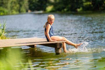 Young girl smiling to camera and kicking water while sitting on a wooden dock by a lake on a bright day at summer- Stock Photo or Stock Video of rcfotostock | RC Photo Stock