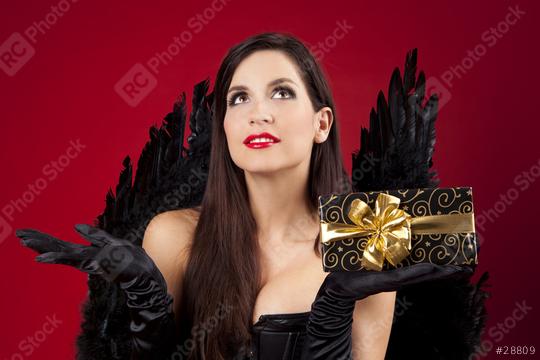 A glamorous woman dressed in black with feathered wings holds a beautifully wrapped gift with a golden bow, set against a bold red background  : Stock Photo or Stock Video Download rcfotostock photos, images and assets rcfotostock | RC Photo Stock.: