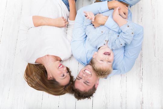 A joyful family of three lies on a white wooden floor, smiling and laughing together, with the child playfully interacting between the parents in a heartwarming and fun moment.  : Stock Photo or Stock Video Download rcfotostock photos, images and assets rcfotostock | RC Photo Stock.: