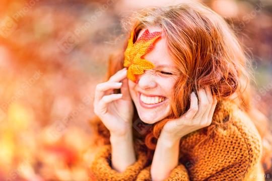 A joyful red-haired woman in an autumn setting, smiling with a vibrant orange leaf covering one eye, wearing a cozy sweater and surrounded by warm fall tones and sunlight  : Stock Photo or Stock Video Download rcfotostock photos, images and assets rcfotostock | RC Photo Stock.: