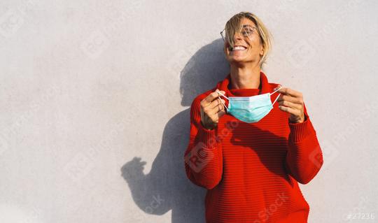 A joyful woman in a bright red sweater removing a light blue face mask, standing against a neutral background, with her shadow visible. Mask freedom after Corona pandemic concept image   : Stock Photo or Stock Video Download rcfotostock photos, images and assets rcfotostock | RC Photo Stock.: