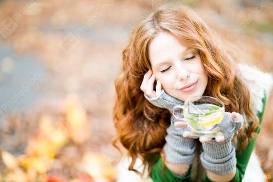 A serene woman with red hair, closed eyes, and a peaceful smile holding a cup of lemon mint tea, sitting outdoors in autumn with colorful leaves in the background, enjoying a cozy moment  : Stock Photo or Stock Video Download rcfotostock photos, images and assets rcfotostock | RC Photo Stock.: