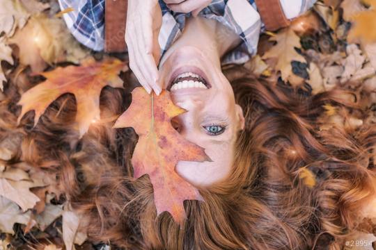 A smiling red-haired woman lies among autumn leaves, playfully holding a large orange leaf near her face, with her vibrant hair spread out, capturing the essence of fall.  : Stock Photo or Stock Video Download rcfotostock photos, images and assets rcfotostock | RC Photo Stock.: