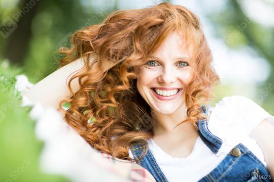 A smiling red-haired woman with curly hair enjoying a sunny outdoor moment, wearing denim overalls and lying on the grass, exuding joy and natural beauty with a vibrant background  : Stock Photo or Stock Video Download rcfotostock photos, images and assets rcfotostock | RC Photo Stock.: