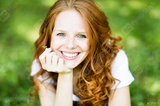A smiling young woman with red curly hair, bright eyes, and freckles lying on grass, holding a small leaf, radiating happiness, natural beauty, and a connection to nature in a sunny park  : Stock Photo or Stock Video Download rcfotostock photos, images and assets rcfotostock | RC Photo Stock.: