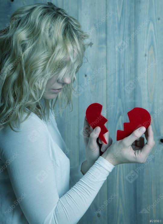 A woman with blonde hair looks down while holding two pieces of a broken red heart against a wooden backdrop, evoking feelings of sadness and heartbreak.  : Stock Photo or Stock Video Download rcfotostock photos, images and assets rcfotostock | RC Photo Stock.: