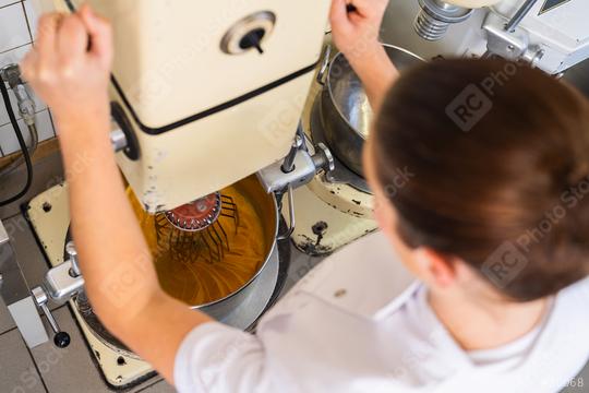 baker operates a commercial stand mixer, mixing a batter or dough. The mixing bowl is filled with a yellow-orange mixture, and the baker is adjusting the machine