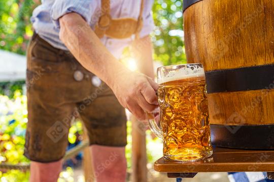 Bavarian man in traditional leather trousers puts fresh poured lager lager beer on the table in front of a wooden beer barrel in the beer garden or oktoberfest  : Stock Photo or Stock Video Download rcfotostock photos, images and assets rcfotostock | RC Photo Stock.: