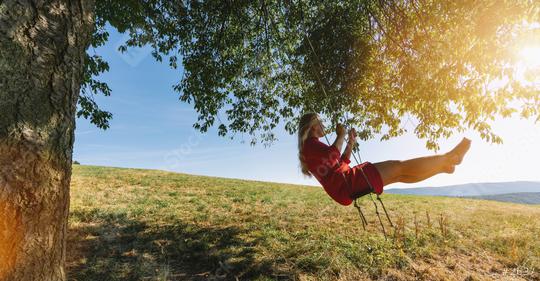 Cheerful lady in red sundress leaning back in her swing in sunny golden countryside. Evening sunshine beaming on lovely girl swaying under single tree in big green meadow  : Stock Photo or Stock Video Download rcfotostock photos, images and assets rcfotostock | RC Photo Stock.: