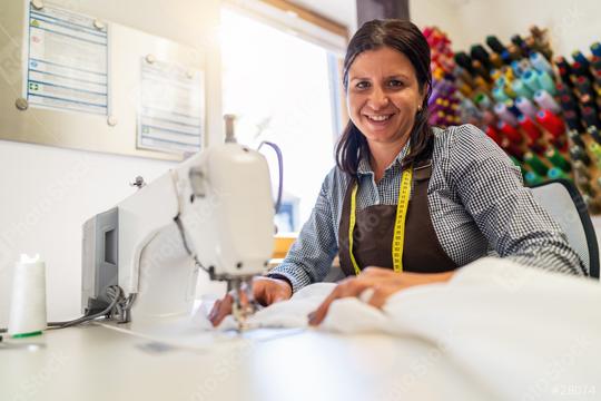 cheerful needlewoman woman with a tape measure around her neck is sitting by a sewing machine in a brightly lit workshop, holding a white silk fabric with colorful spools of thread in the background  : Stock Photo or Stock Video Download rcfotostock photos, images and assets rcfotostock | RC Photo Stock.: