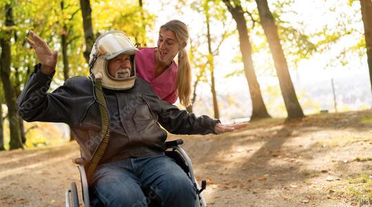 Elderly man in an astronaut helmet extends his arms in a flying gesture, with a nurse smiling beside him in a sunlit autumn park. Dementia retirement home concept image  : Stock Photo or Stock Video Download rcfotostock photos, images and assets rcfotostock | RC Photo Stock.: