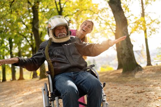 Elderly man in an astronaut helmet joyfully extends arms, imitating flight, with a nurse smiling behind him in a sunny park. Dementia retirement home concept image  : Stock Photo or Stock Video Download rcfotostock photos, images and assets rcfotostock | RC Photo Stock.: