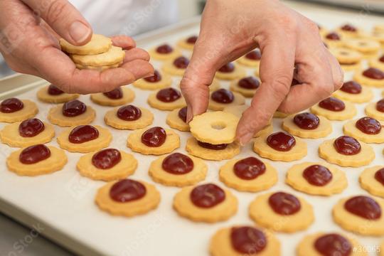 Hands assembling Linzer cookies by placing a top layer with a hole over a bottom cookie with a dollop of red jam. Rows of prepared cookies lie on a baking sheet in the background  : Stock Photo or Stock Video Download rcfotostock photos, images and assets rcfotostock | RC Photo Stock.: