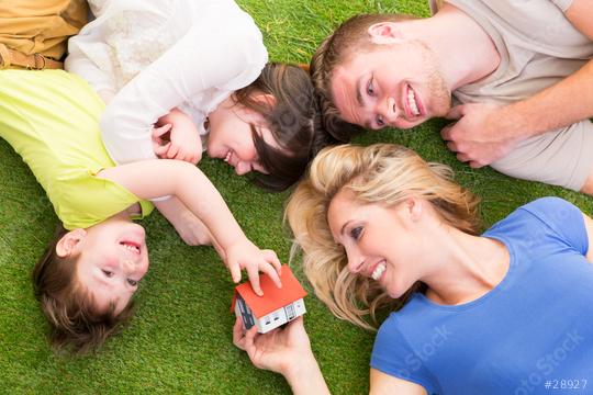 Happy family of four lying on green grass, smiling and playing with a miniature house, symbolizing home ownership, family bonding, and togetherness in a bright and cheerful outdoor setting  : Stock Photo or Stock Video Download rcfotostock photos, images and assets rcfotostock | RC Photo Stock.: