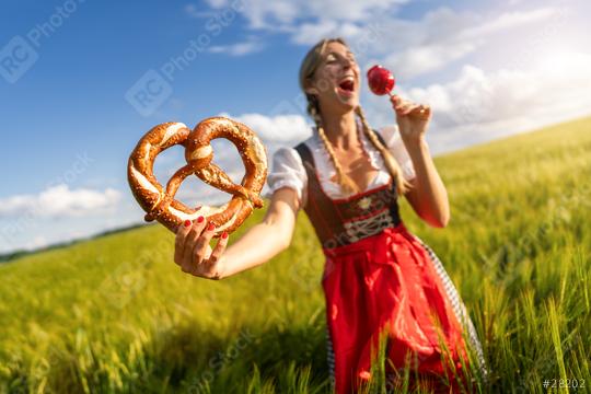 Joyful woman in Bavarian dress holding a pretzel and a candy apple, laughing in a wheat field celebrating Oktoberfest ferstival in munich  : Stock Photo or Stock Video Download rcfotostock photos, images and assets rcfotostock | RC Photo Stock.: