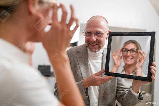 Optician holding a mirror to smiling customer trying on new glasses in optical store.  : Stock Photo or Stock Video Download rcfotostock photos, images and assets rcfotostock | RC Photo Stock.: