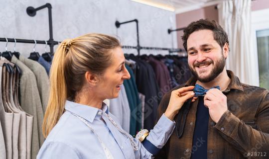 Saleswoman fitting a bow tie on a customer, both are smiling, racks of clothes behind in a men