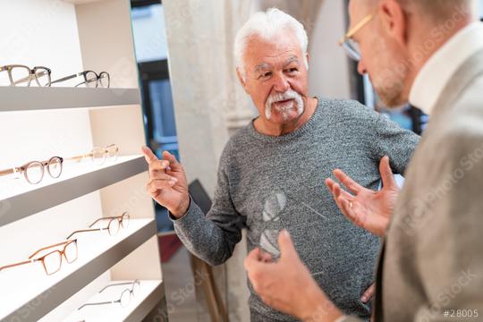 Senior man gesturing during a conversation with an optician in an optical shop with glasses displayed in the background  : Stock Photo or Stock Video Download rcfotostock photos, images and assets rcfotostock | RC Photo Stock.: