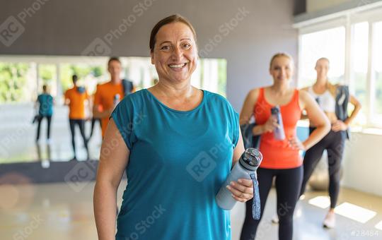 Smiling older woman holding a water bottle in a gym with other participants in the background. Teamwork Concept image  : Stock Photo or Stock Video Download rcfotostock photos, images and assets rcfotostock | RC Photo Stock.: