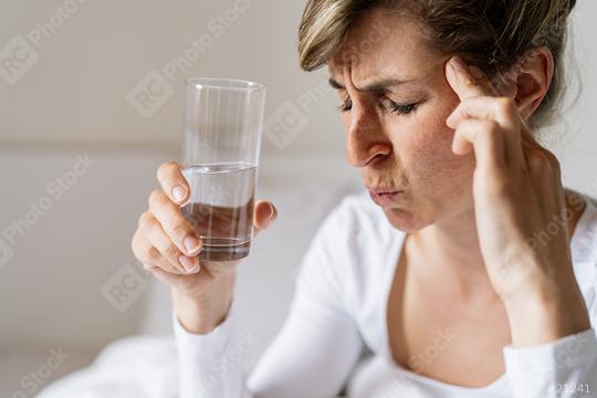 tired woman touching forehead and holding glass with water having headache migraine or depression, feel stressed grabs head with hand with pain sit on bed in the morning, Flu or overload Concept   : Stock Photo or Stock Video Download rcfotostock photos, images and assets rcfotostock | RC Photo Stock.: