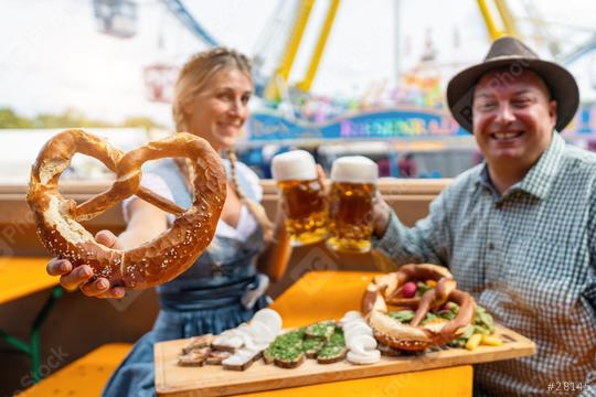 Woman holding a pretzel and toasting beer mugs with a man at a beer tent table with snacks on a wooden board and amusement rides at oktoberfest or dult in germany  : Stock Photo or Stock Video Download rcfotostock photos, images and assets rcfotostock | RC Photo Stock.: