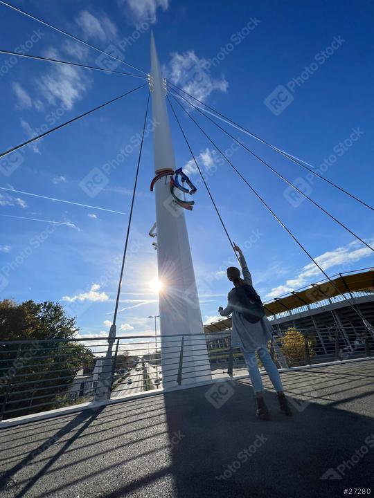 Woman in a gray cardigan and jeans stands on a bridge, reaching up towards a colorful horse emblem on a pole, with a bright sun, blue sky, and modern building in the background
  : Stock Photo or Stock Video Download rcfotostock photos, images and assets rcfotostock | RC Photo Stock.: