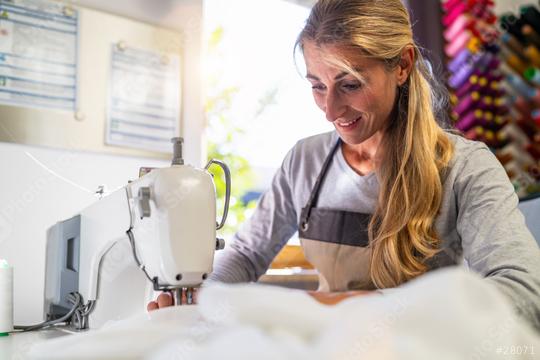 woman in an apron is working at a sewing machine, stitching white fabric in a well-lit workspace with colorful thread spools in the background  : Stock Photo or Stock Video Download rcfotostock photos, images and assets rcfotostock | RC Photo Stock.: