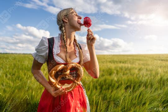 Woman in Bavarian tracht tasting a candy apple and holding a pretzel in a sunny wheat field celebrating Oktoberfest in munich  : Stock Photo or Stock Video Download rcfotostock photos, images and assets rcfotostock | RC Photo Stock.: