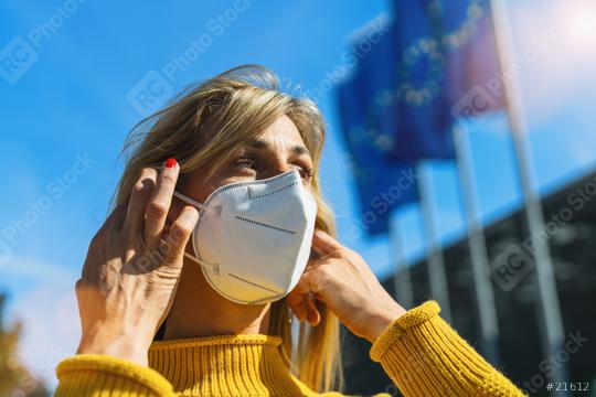 Woman in the city puts on a medical anti virus protection knN95 FFP2 face mask to prevent others from corona virus COVID-19 and SARS cov 2 infection with european flags  in the background  : Stock Photo or Stock Video Download rcfotostock photos, images and assets rcfotostock | RC Photo Stock.: