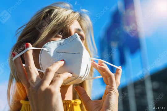 Woman in the city puts on a medical anti virus protection knN95 FFP2 face mask to prevent others from corona virus COVID-19 and SARS cov 2 infection with european flags  in the background  : Stock Photo or Stock Video Download rcfotostock photos, images and assets rcfotostock | RC Photo Stock.: