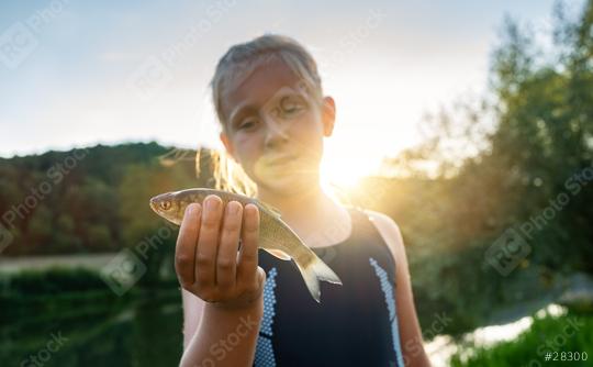 Young girl proudly displaying a small fish in her hand against a backdrop of sunset and lush greenery  : Stock Photo or Stock Video Download rcfotostock photos, images and assets rcfotostock | RC Photo Stock.: